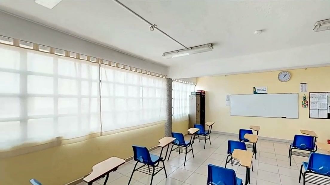 Aula con pupitres individuales azules, ventanales con cortinas y pizarrón blanco.
