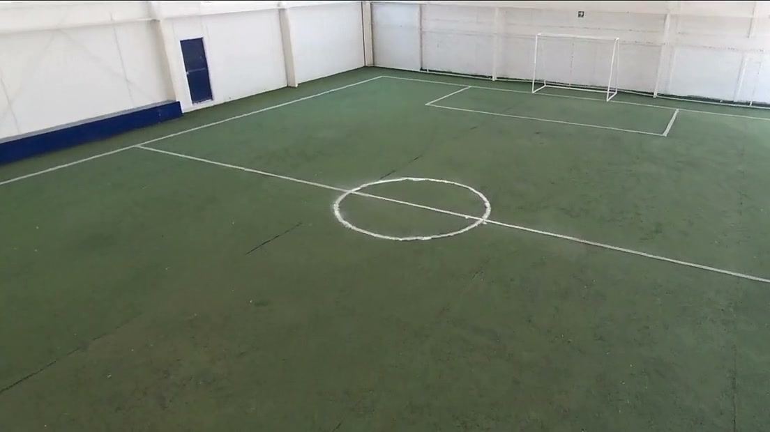Cancha de pasto sintético verde con líneas blancas, círculo central y porterías, rodeada de muros blancos.